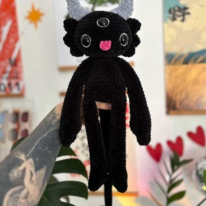 May include: A black crocheted plush creature featuring three eyes, two light gray horns, and a small pink tongue. The figure has long arms, a long tail ending in a light gray heart-shaped tip, and a textured, soft yarn construction throughout.
