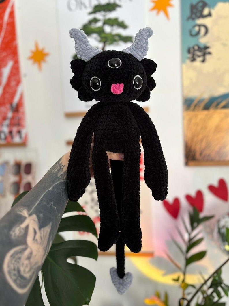May include: A black crocheted plush creature featuring three eyes, two light gray horns, and a small pink tongue. The figure has long arms, a long tail ending in a light gray heart-shaped tip, and a textured, soft yarn construction throughout.