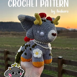 May include: A crocheted plush creature with grey fur, yellow antlers, and red accents. The plushie has a white flower and pearl necklace. The text "CROCHET PATTERN by Amikoro" is displayed at the top of the image.