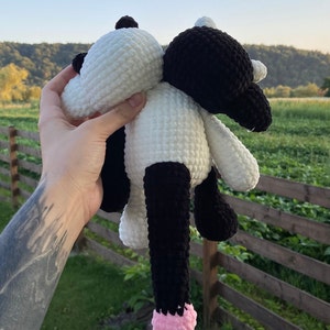 May include: A handmade crochet cow plushie in black and white. The plush toy has a white head and body, black legs and ears, and a pink tail. The cow is held up against a background of green grass and trees.