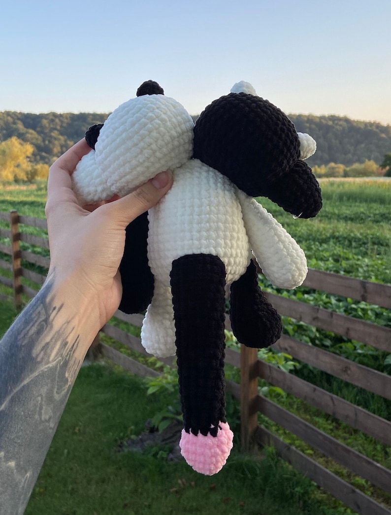 May include: A handmade crochet cow plushie in black and white. The plush toy has a white head and body, black legs and ears, and a pink tail. The cow is held up against a background of green grass and trees.
