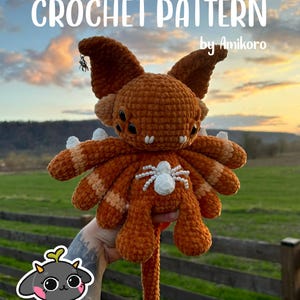 Könnte beinhalten: Eine gehäkelte orange-weiß Kreatur mit mehreren Augen, großen Ohren und einem langen Schwanz. Der Text "CROCHET PATTERN by Amikoro" ist oben zu sehen. Eine kleine Spinne ist am Ohr der Kreatur befestigt. Der Hintergrund ist ein Sonnenuntergang.