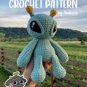 May include: A crocheted alien plushie in shades of teal, with large black eyes, star-shaped pupils, and yellow antennae. The text "CROCHET PATTERN" and "by Amikoro" are displayed at the top. The plushie is held in front of a blurred outdoor background.