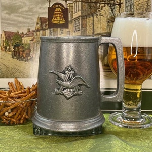 May include: A pewter beer stein with a silver eagle logo and a vintage postcard image of a pub in the background. The stein has a handle and a wide base.