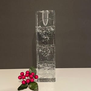 May include: A clear, rectangular glass candle holder with a textured surface. The candle holder is filled with bubbles and has a small opening at the top. A sprig of red berries with green leaves sits in front of the holder.