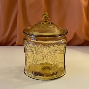 May include: Amber-colored glass jar with a lid. The jar has a square shape with rounded corners and embossed decorative patterns. The lid has a small knob on top. The jar is sitting on a white surface.