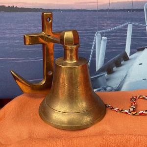 May include: A brass ship's bell with a clapper and a matching brass anchor. The bell is sitting on an orange cloth. A multi-colored rope is attached to the bell. The background shows a nautical scene.