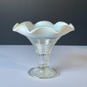 May include: A clear glass sundae dish with a ruffled, opalescent rim. The bowl transitions from white to a pale blue, supported by a ribbed stem and a circular base. The dish is designed for serving desserts.
