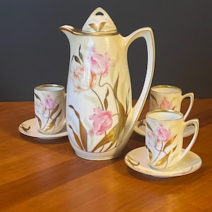 May include: A vintage porcelain tea set featuring a cream-colored teapot and four matching cups and saucers. The set is decorated with pink tulip floral designs and gold accents. The teapot has a tall, slender shape with a lid.