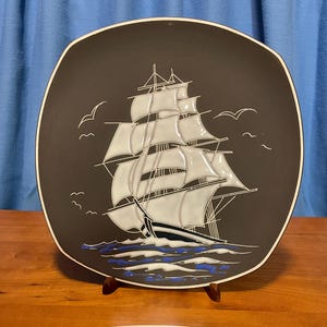 May include: Square black plate with a white rim, featuring a detailed illustration of a sailing ship with white sails and black accents. The ship sails on blue waves, with white seagulls in the sky.