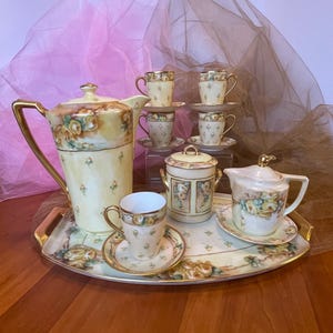 May include: A vintage tea set with a cream and gold color scheme. The set includes a teapot, sugar bowl, creamer, cups, and saucers, all decorated with floral patterns. The set is arranged on a decorative tray.