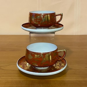 May include: Two matching teacups and saucers. The cups and saucers are a deep red with gold bird and floral designs. The rims of the saucers are white, and the cup handles are gold. The cups have a white interior.