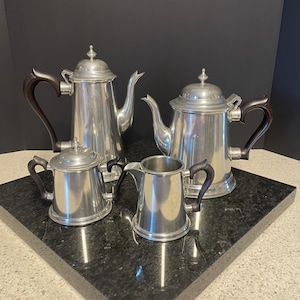 May include: A four-piece silver-toned metal tea set consisting of two tall teapots with lids, a lidded sugar bowl, and a creamer. Each piece features a smooth, polished finish and dark brown curved handles, with a finial on each lid.