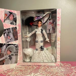 Puede incluir: Muñeca en caja, vestida con un vestido blanco con detalles negros, un sombrero grande y una sombrilla. La caja presenta imágenes de una mujer con un atuendo similar. Es un artículo de colección.