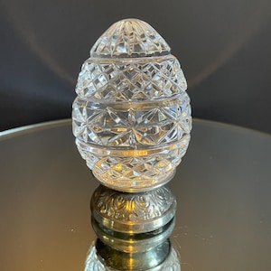 May include: A clear, egg-shaped crystal jar with a diamond-cut pattern. The jar has a silver-colored base and lid. The jar is sitting on a reflective surface.