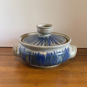 May include: A ceramic casserole dish with a lid. The dish is off-white with blue brushstroke designs. It has two handles and a small knob on the lid. The dish is sitting on a wooden surface.