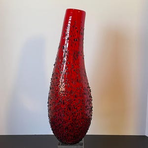 May include: A tall, red glass vase with a textured surface and black speckles. The vase has a unique, elongated shape and sits on a clear, square base. The vase is a decorative art piece.