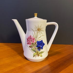 May include: White ceramic coffee pot with a floral design featuring pink, blue, and white flowers. The pot has a long spout, a handle, and a gold-colored lid finial. The flowers are painted with green leaves.