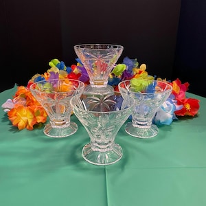 May include: Four clear glass dessert bowls with intricate starburst and leaf patterns. The bowls are arranged on a green surface, surrounded by colorful leis. The background is black.