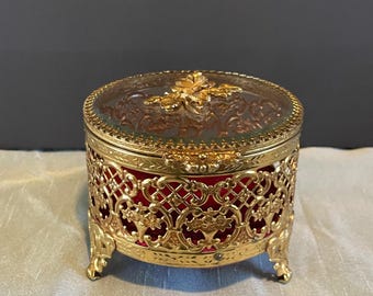 May include: A round gold-tone metal trinket box with an intricate filigree pattern and three small feet. The lid features a clear glass top with a gold-tone floral accent in the center. The interior is lined with a vibrant red velvet fabric.