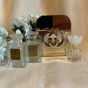 May include: A collection of perfume bottles, including a gold-toned Gucci Guilty bottle and several small, clear glass bottles with labels. The bottles are arranged on a light-colored surface with white flowers in the background.