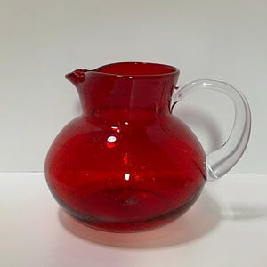 May include: A red glass pitcher with a clear glass handle. The pitcher has a rounded body and a spout for pouring. The glass has a textured appearance. The pitcher is likely used for serving beverages.