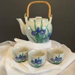 May include: A ceramic teapot with a woven rattan handle and three matching small cups. The off-white glazed set features hand-painted blue iris flowers with green stems and leaves. The teapot includes a lid and a curved spout for pouring.