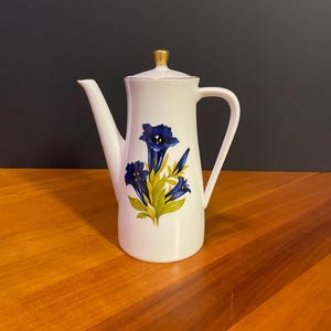 May include: White ceramic coffee pot with a blue floral design and a gold-colored lid. The pot has a long spout and a handle. The floral design features blue flowers with green leaves. The pot is on a wooden surface.
