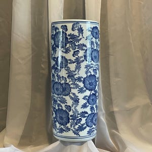 May include: A cylindrical ceramic vase featuring a white base with a repeating blue floral pattern. The design consists of detailed blossoms and leafy foliage covering the entire exterior surface, finished with a smooth, glossy glaze.