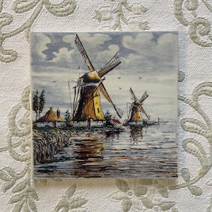 May include: A vintage tile with a hand-painted image of two Dutch windmills in a landscape setting. The windmills are yellow and white with blue blades. The sky is a light blue with white clouds. The water is a dark blue with reflections of the windmills.
