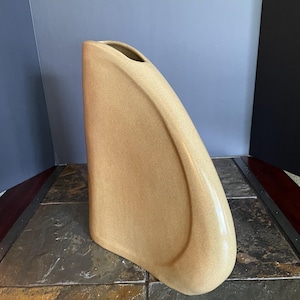 May include: A tall, sculptural ceramic vase with a smooth, matte, light tan finish. The vase features a flat, vertical back edge and a curved, sweeping front profile that tapers toward a narrow, elongated opening at the top center.