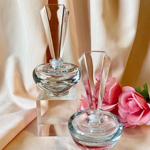 May include: Two clear glass perfume bottles with geometric stoppers. The bottles have a round base with a red tint and sit on a light pink surface. A pink rose is in the background. The bottles are empty.