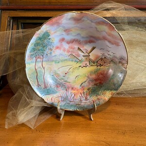 May include: A decorative ceramic plate with a landscape scene featuring a windmill, trees, and a river. The plate has a gold rim and is displayed on a gold-colored stand. The artwork uses a variety of colors, including green, blue, red, and yellow.