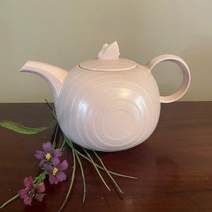 May include: A light pink ceramic teapot with a circular design and a butterfly-shaped handle on the lid. The teapot has a curved spout and a rounded handle. Purple flowers and green stems are arranged on a dark brown surface.