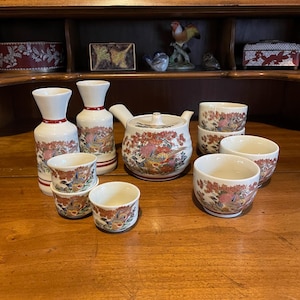 May include: A Japanese tea set with a white background, featuring a teapot, cups, and sake bottles. The set is decorated with a colorful bird and floral design. The set includes a teapot, two sake bottles, and eight cups.