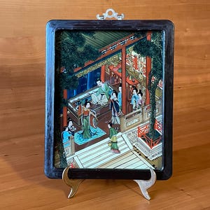 May include: A framed Asian-style painting depicting a courtyard scene with figures in traditional attire. The artwork features vibrant colors, a dark wooden frame, and a decorative metal hanger. The painting is displayed on a gold-toned easel.