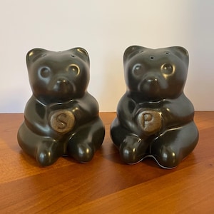 May include: A pair of dark green ceramic bear-shaped salt and pepper shakers. The salt shaker has an "S" on its belly, and the pepper shaker has a "P". Each shaker has three holes on top for dispensing.