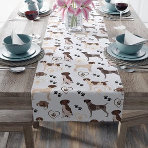 May include: White table runner featuring a repeating pattern of brown and tan dogs, paw prints, and hearts. The runner is set on a wooden table with place settings, wine glasses, and a vase of pink flowers.
