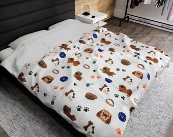 Könnte beinhalten: Weiße Decke mit einem sich wiederholenden Muster aus braunen Hunden, Pfotenabdrücken, Knochen, Hundehalsbändern und blauen und grünen Spielzeugen. Die Decke liegt auf einem Bett mit weißen Laken und einem schwarzen Kopfteil. Die Decke ist aus weichem Material.