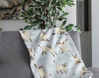 May include: A light blue blanket featuring a pattern of Siamese cats in various poses, with the word "MEOW" and small blue flowers scattered across the surface. The blanket is draped over a gray armchair.