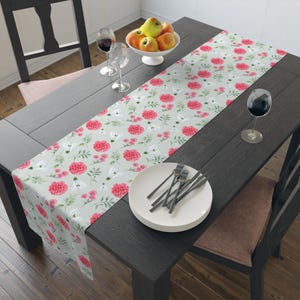 May include: A floral table runner with a light green background and a pattern of pink flowers and white blossoms. The runner is placed on a dark wooden table, set with plates, forks, and wine glasses. A bowl of apples sits nearby.