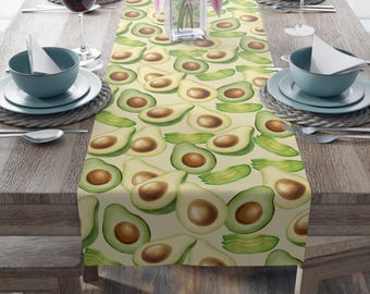May include: A table runner featuring a repeating pattern of avocado halves and slices in shades of green and brown on a light beige background. The runner is placed on a wooden table set for a meal, with plates, glasses, and flowers.