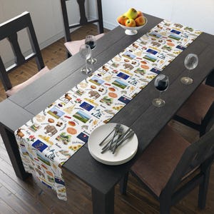 May include: A white table runner with a repeating pattern of illustrations and text. The design includes images of animals, landscapes, and state flags. The runner is placed on a dark wooden table, with place settings and wine glasses.