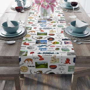 Puede incluir: Un camino de mesa blanco con un patrón repetido de nombres de ciudades e ilustraciones de Ohio. El camino de mesa está sobre una mesa de madera preparada para una comida, con platos, cuencos y copas de vino. Un jarrón de flores rosas está en el centro.