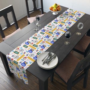 May include: A white table runner with a repeating pattern of school supplies, including rulers, pencils, scissors, and books. The runner also features the numbers "123" and the letter grade "A+". The runner is on a dark wooden table.