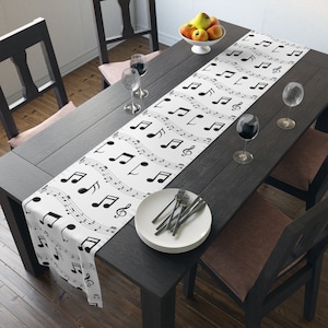 May include: White table runner with a black musical note design. The runner is on a dark wood table with two chairs and a bowl of fruit.