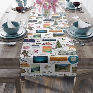 May include: A white table runner with a repeating pattern of South Dakota imagery, including illustrations of landmarks, towns, and wildlife. The runner is set on a wooden table with place settings and a floral centerpiece.