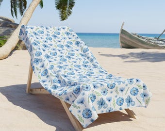 Może przedstawiać: Białe leżak plażowy z niebiesko-zielonym ręcznikiem w kwiatowy wzór. Leżak stoi na piaszczystej plaży z drewnianym łódką w tle.