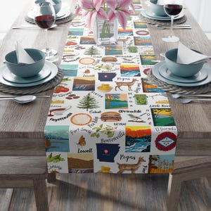 Op de afbeelding: Een witte tafelloper met kleurrijke vierkanten met illustraties en tekst van steden en bezienswaardigheden in Arkansas. De loper ligt op een houten tafel die gedekt is voor een maaltijd, met borden, kommen en wijnglazen.