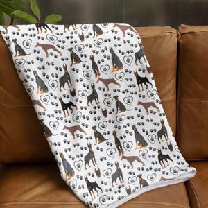 May include: White blanket featuring a repeating pattern of Doberman Pinschers in various poses, along with paw prints and heart shapes. The blanket is folded over a brown leather couch, showcasing the design's details.